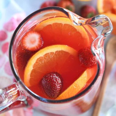 Top down view of red punch in glass pitcher with sliced oranges and strawberries.
