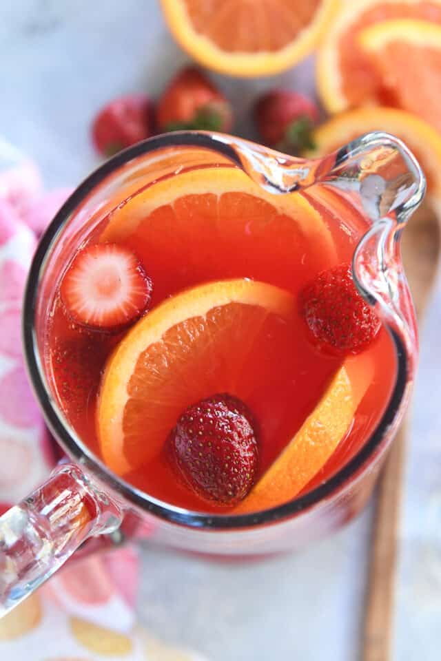 Top down view of red punch in glass pitcher with sliced oranges and strawberries.