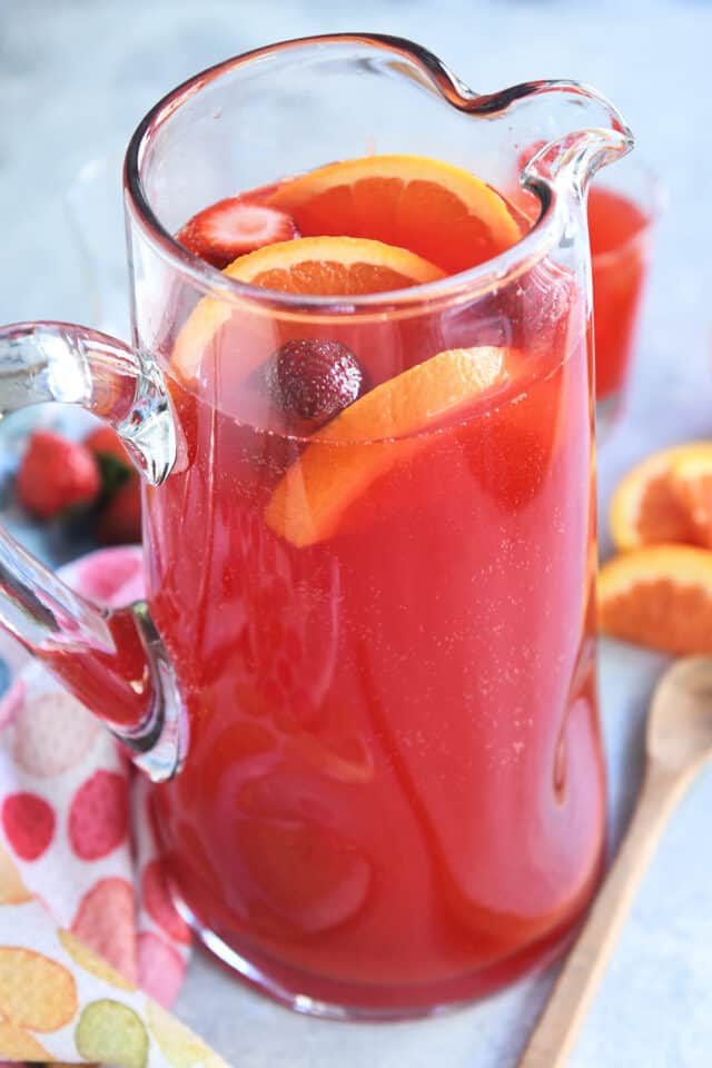 Red punch in glass pitcher with sliced oranges and strawberries.