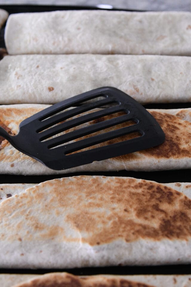 Pressing beef and bean tortilla wrap on griddle with spatula.