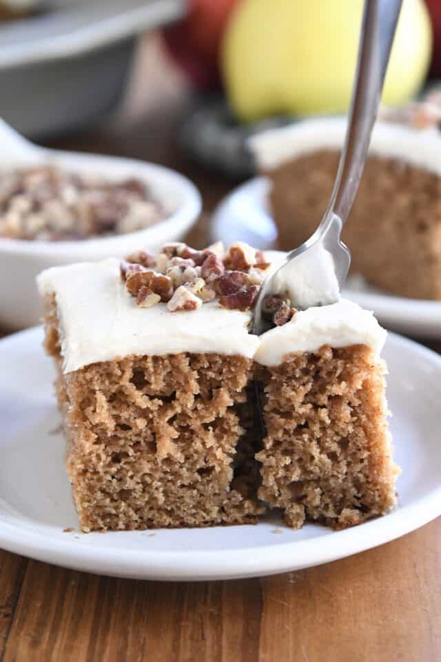 Fork taking bite of frosted applesauce spice cake on white plate.