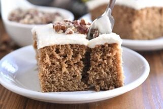 Fork taking bite of frosted applesauce spice cake on white plate.