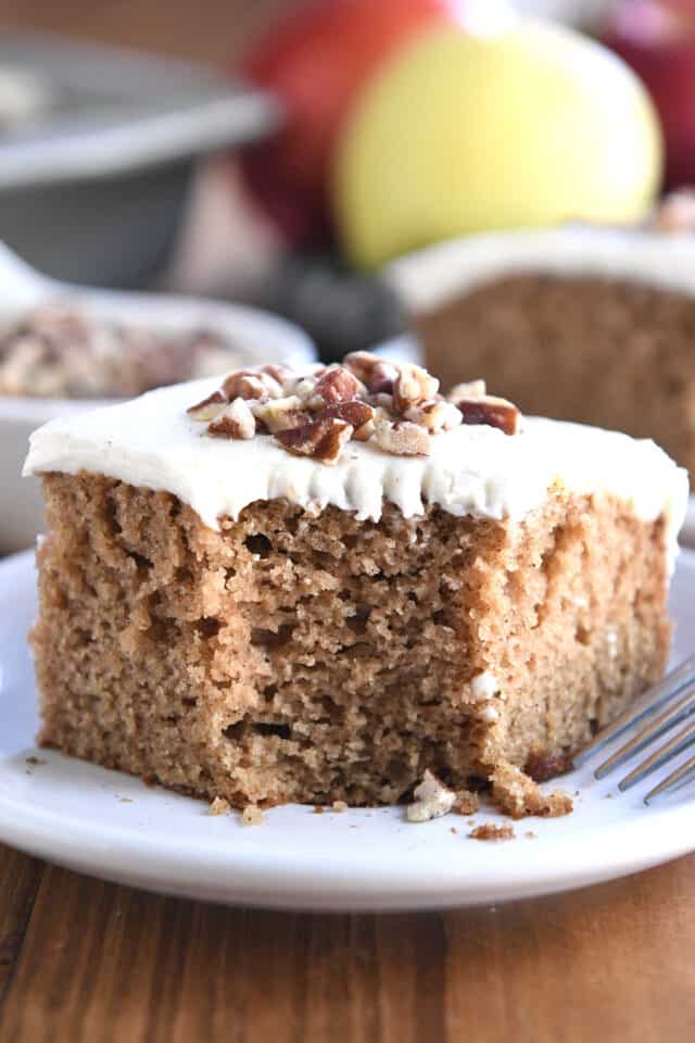 Square of frosted applesauce spice cake on white plate with pecans on top.
