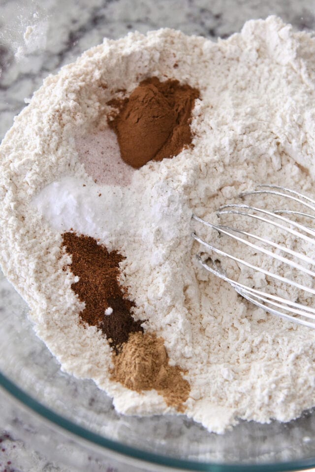 Glass bowl with whisk stirring flour, cinnamon, allspice, cloves, ginger, salt and baking soda.