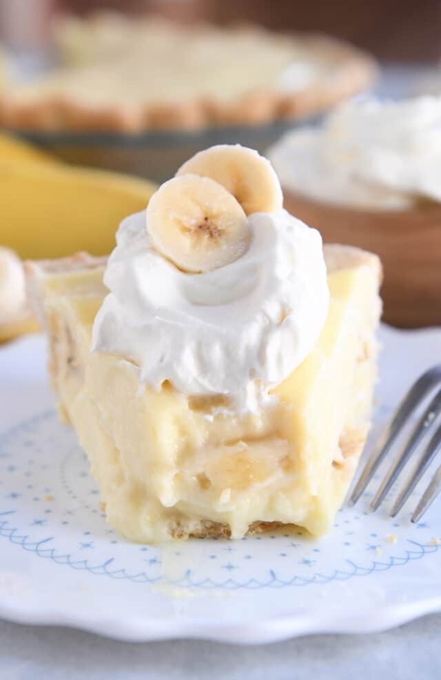 Bite taken out of piece of banana cream pie on fluted plate topped with whipped cream and sliced bananas.