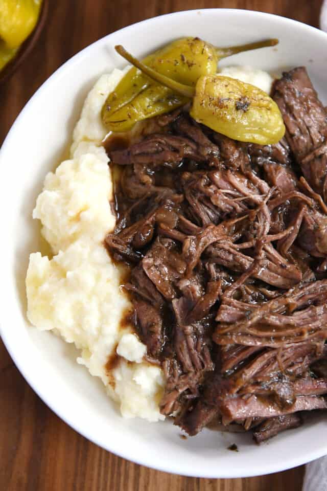 Top down view of white shallow bowl with mashed potatoes, shredded beef and gravy, and two pepperoncini.