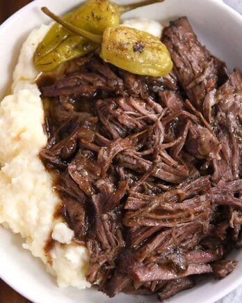 White shallow bowl with mashed potatoes, two pepperoncini, and shredded beef.