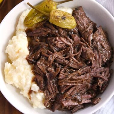 White shallow bowl with mashed potatoes, two pepperoncini, and shredded beef.