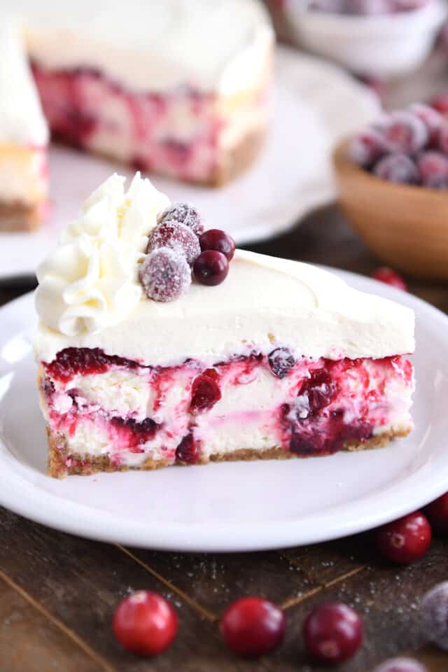 Slice of cranberry white chocolate mousse cheesecake on white plate with piped whipped cream and sugared cranberries.