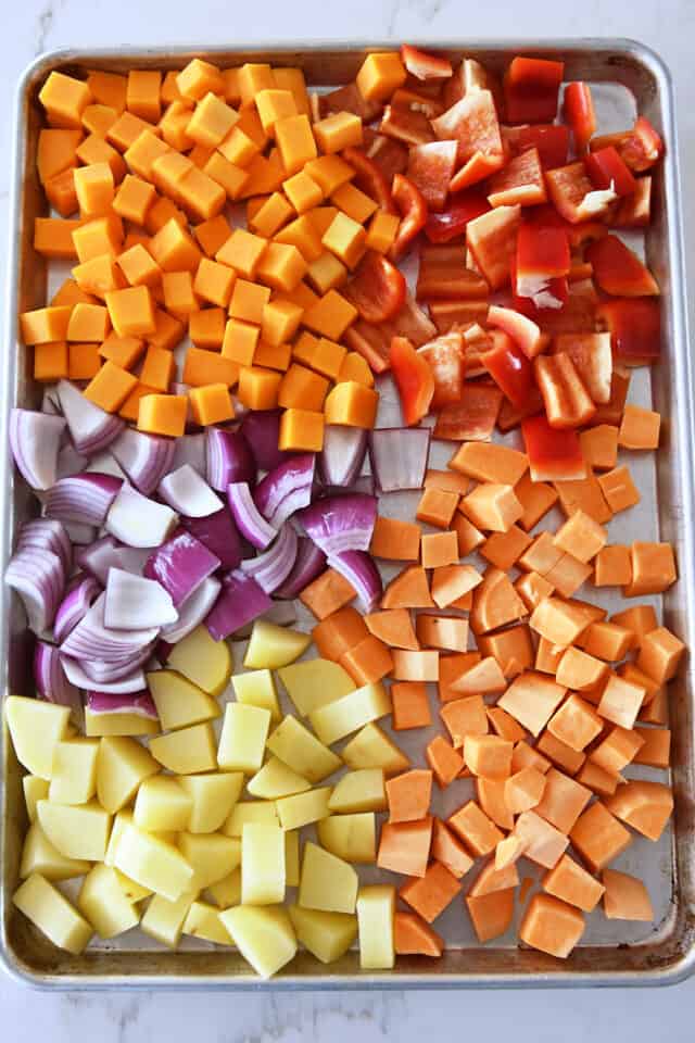 Sheet pan with uncooked butternut squash, sweet potatoes, red bell peppers, red onion and potatoes.