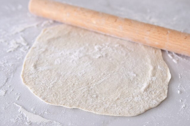 Rolling flatbread dough into thin circle on floured counter.