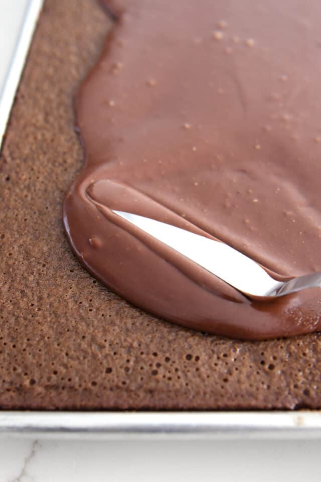 Using offset spatula to spread thin frosting over chocolate cake in sheet pan.
