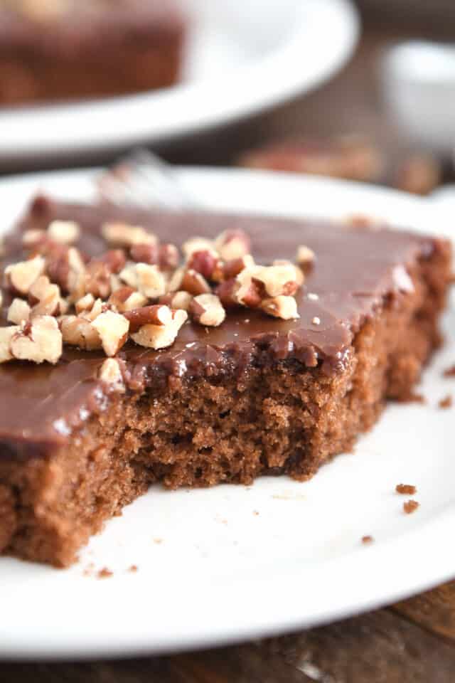 Piece removed from square of chocolate Texas sheet cake on white plate with pecans on top.