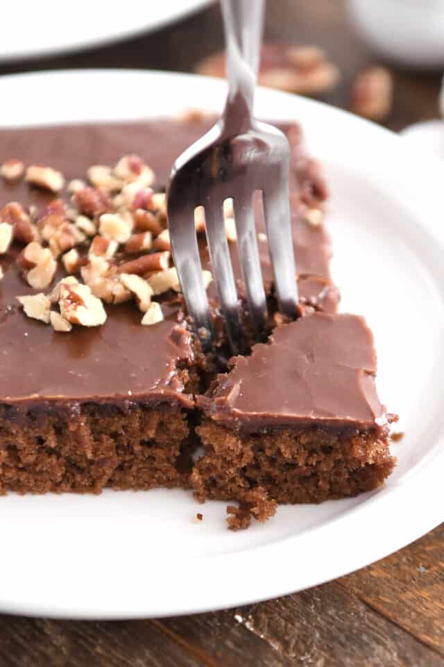 Fork taking corner piece from serving of frosted chocolate cake on white plate.