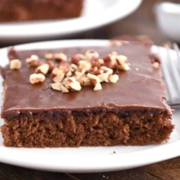 Square of frosted chocolate Texas sheet cake on white plate with pecans on top.