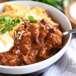 Red chili with spoonful in gray bowl with sour cream, green onions, and crushed corn chips.