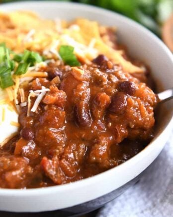 Red chili with spoonful in gray bowl with sour cream, green onions, and crushed corn chips.