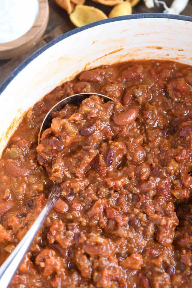 Red chili with ground beef and beans in white cast iron pot with ladle.