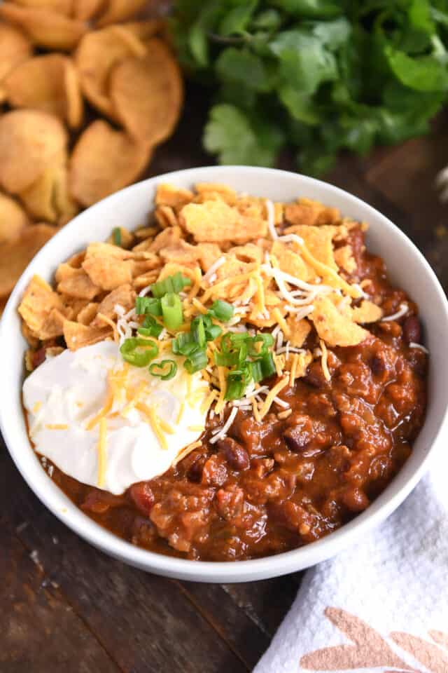 Red chili with beans and ground beef in gray bowl topped with sour cream, shredded cheese, green onions, and crushed corn chips.