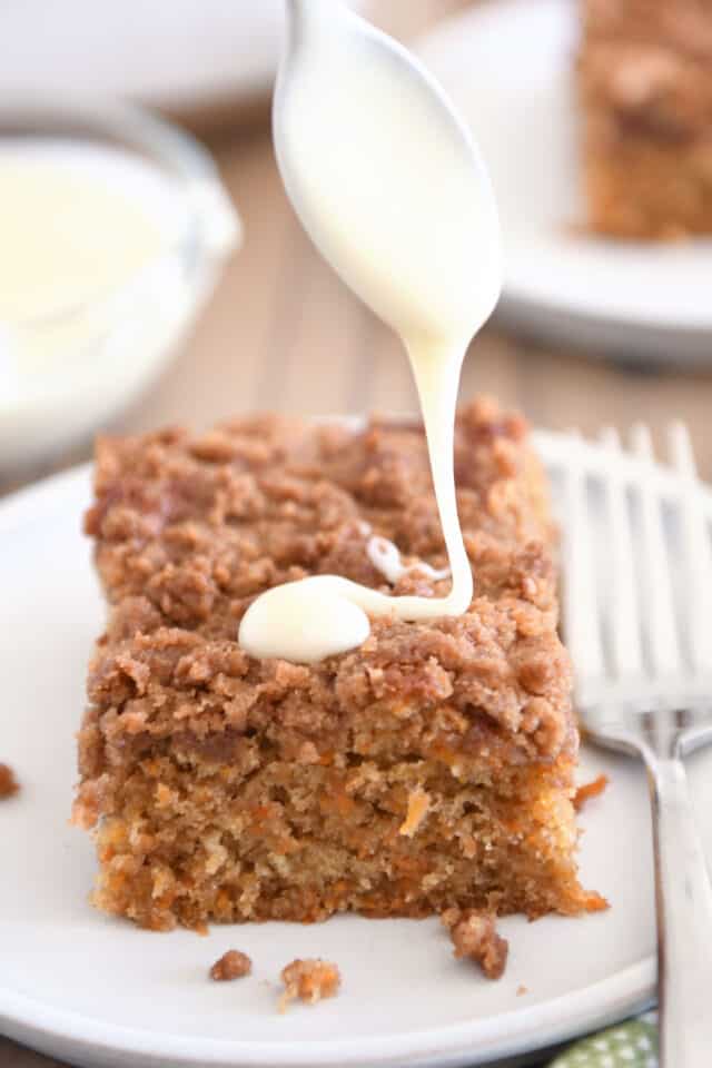 Drizzling cream cheese glaze over square of carrot cake coffee cake on white plate.