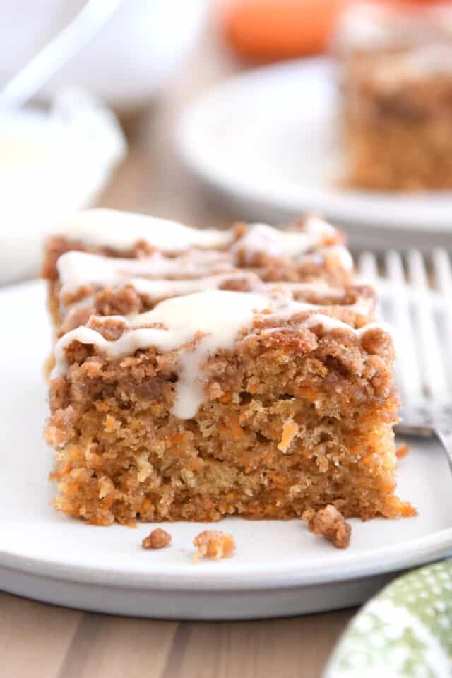 Square of carrot cake coffee cake drizzled with cream cheese glaze on white plate with fork.