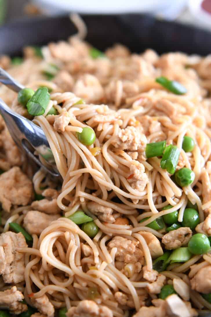 Tongs grabbing scoop of ramen noodles with peas and green onions.