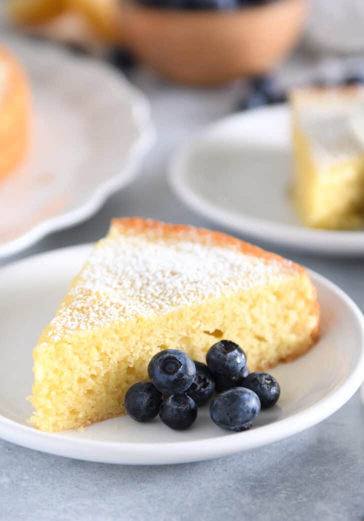 Triangle of olive oil cake dusted with powdered sugar on white plate with blueberries.