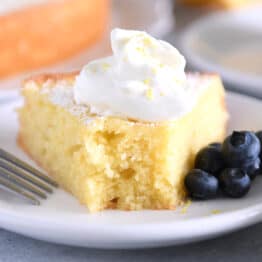 Triangle of olive oil cake with bite taken out with whipped cream and blueberries on white plate.