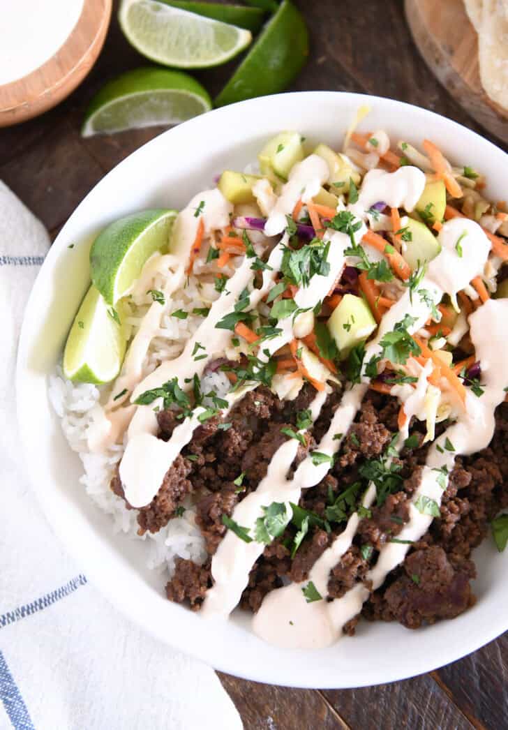 Top down view of white bowl with rice, Korean beef, slaw, cucumbers, sauce and lime wedges.