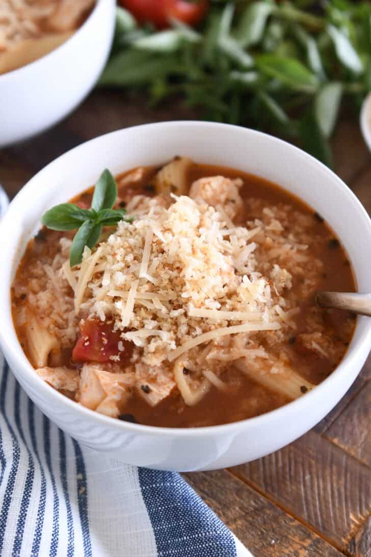 Chicken parmesan soup with cooked pasta, chicken, tomatoes, and seasonings in white bowl topped with buttered bread crumbs with Parmesan.