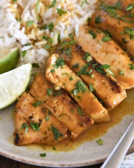 Grilled chicken breast slices with sauce, rice and fresh limes on gray plate.