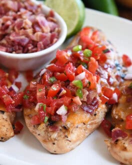 Grilled chicken breasts topped with pico de gallo on a white plate with a bowl of chopped bacon and a half of a lime in the background.