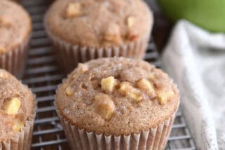 Several apple cinnamon muffins on cooling rack with apples in the background.