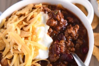 Spoon scooping bite of beef and bean tomato chili in sour cream, shredded cheese and corn chips.