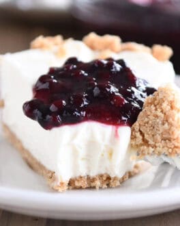 A slice of cheesecake with a dollop of blueberry sauce on top, on a white plate. A fork has a bite of the cheesecake on it next to the slice.