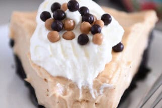 A slice of chocolate cookie crust peanut butter pie with a bite taken out on a white plate. The pie is topped with whipped cream and small chocolate balls.