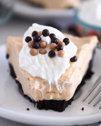 A slice of chocolate cookie crust peanut butter pie with a bite taken out on a white plate. The pie is topped with whipped cream and small chocolate balls.