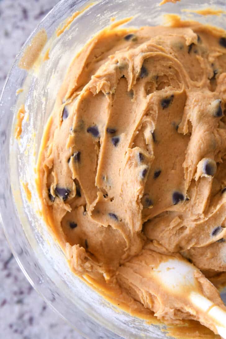 Top down view of a glass bowl filled with pumpkin chocolate chip cookie dough batter with a white rubber spatula on the side of the bowl.