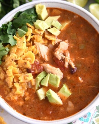Top down view of a white bowl full of chicken tortilla soup topped with diced avocados, crushed chips, and chopped cilantro.