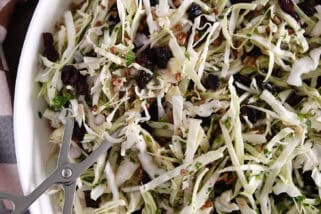 White bowl with salad tongs scooping portion of thin sliced green cabbage, dried cranberries, pecans and red onions.