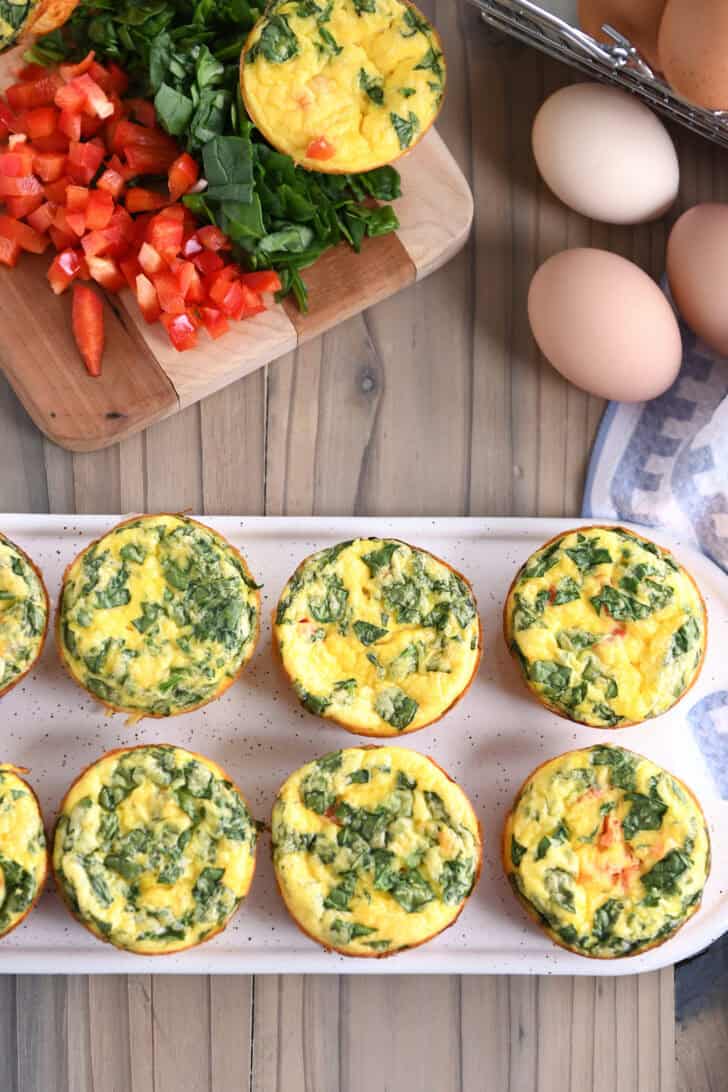 Top down view of cooked egg and veggie muffins cups on a white platter with fresh chicken eggs and a cutting board of chopped vegetables in the background.