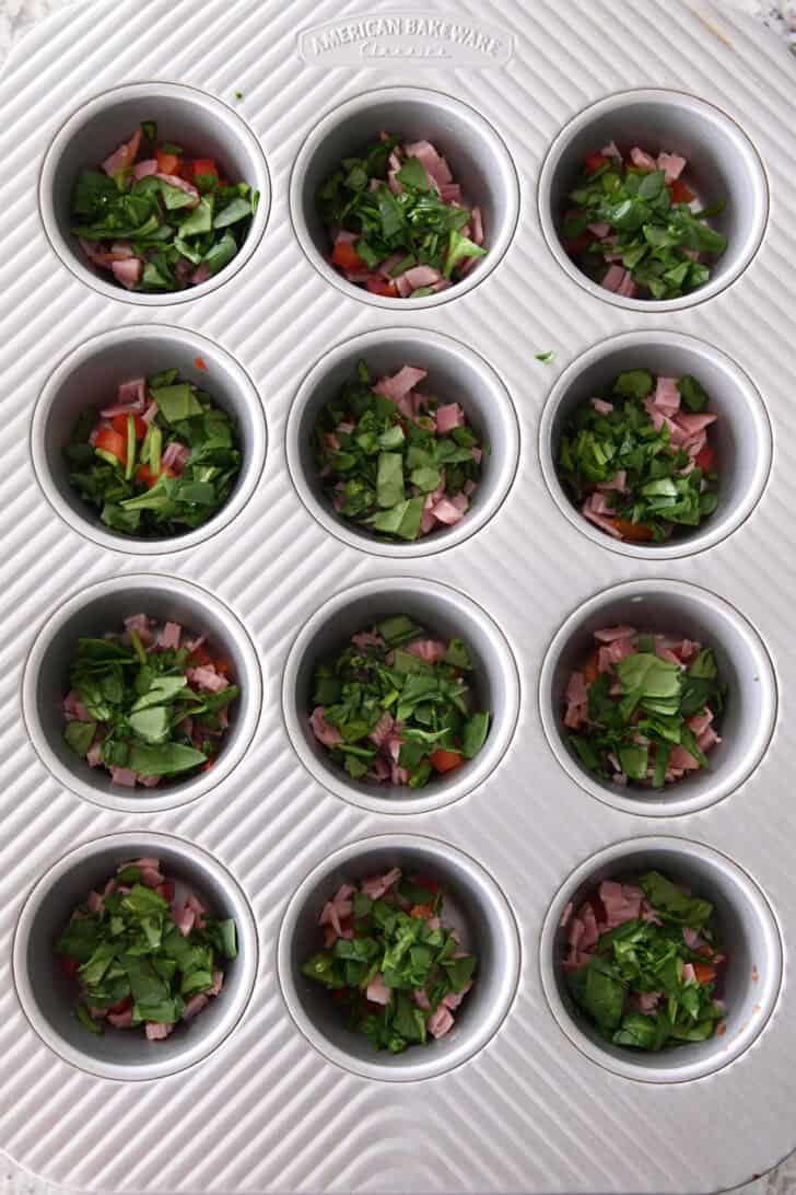 Top down view of a muffin tin, each cup is filled with chopped vegetables and diced ham.