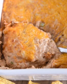 Serving spoon scooping serving of warm bean dip topped with cheese in white square baking dish.
