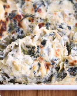 A large spoonful of creamy spinach artichoke dip being taken out of a white casserole dish full of the dip.