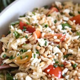 A wooden spoon taking a scoop of cooked orzo salad with tomatoes, pine nuts, and basil out of a white casserole dish.