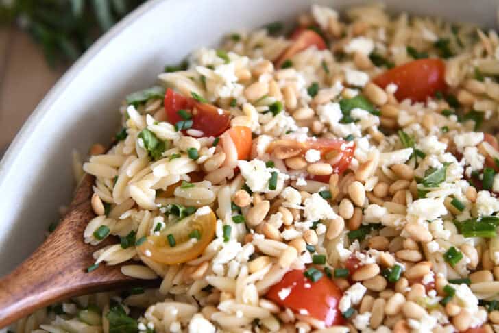 A wooden spoon taking a scoop of cooked orzo salad with tomatoes, pine nuts, and basil out of a white casserole dish.