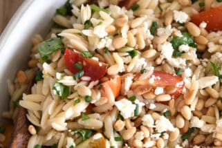 A wooden spoon taking a scoop of cooked orzo salad with tomatoes, pine nuts, and basil out of a white casserole dish.