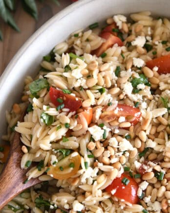A wooden spoon taking a scoop of cooked orzo salad with tomatoes, pine nuts, and basil out of a white casserole dish.