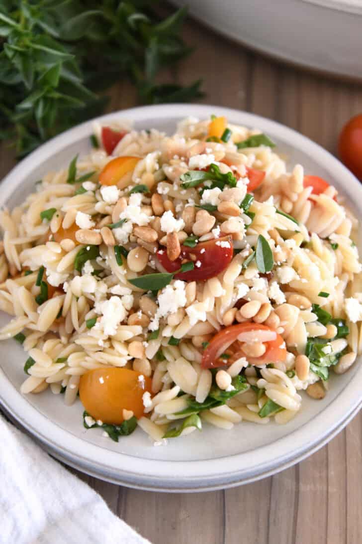 A white plate with cooked orzo, halved cherry tomatoes, chopped basil, pine nuts, and crumbled feta.