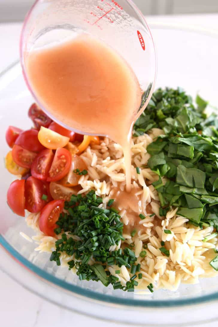 A glass bowl with cooked orzo, chopped scallions, halved cherry tomatoes, and chopped basil. Sauce is being poured over the salad ingredients from a measuring cup.